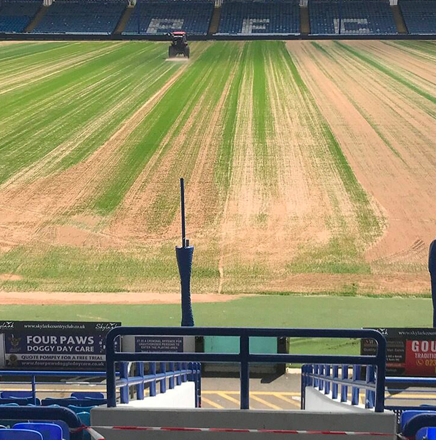 A professional football pitch undergoing deep scarification and renovation works inside a league stadium, with a tractor visible at the far end.