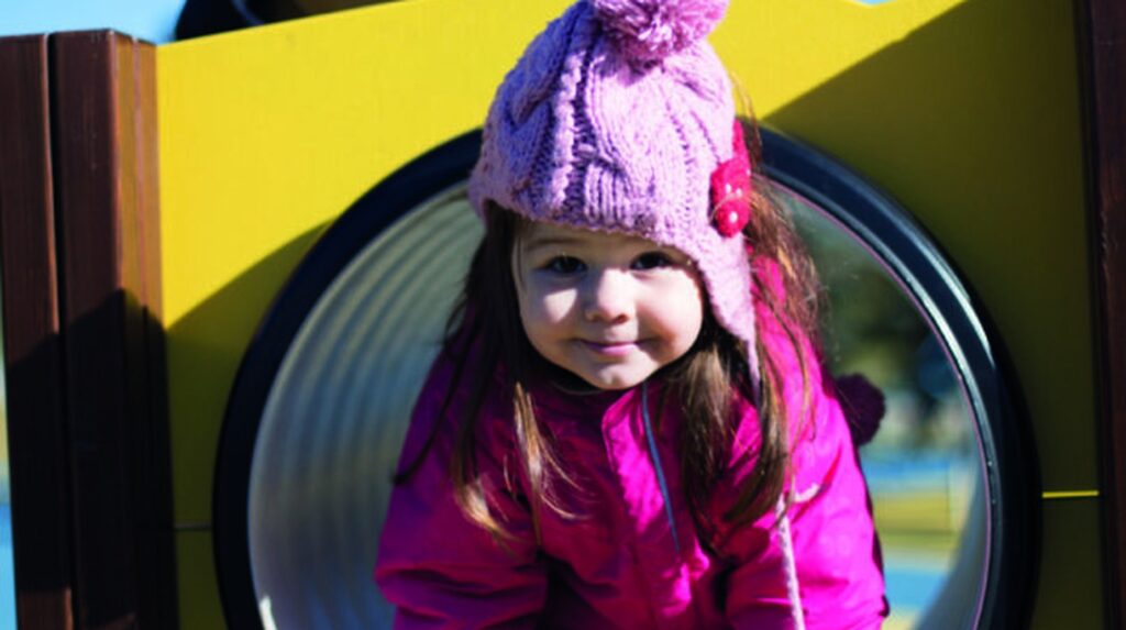 Young girl smiling in playground tunnel wearing pink coat and knit hat