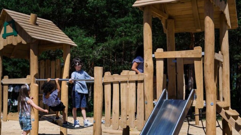 Children playing on a wooden playground structure with slide, surrounded by trees