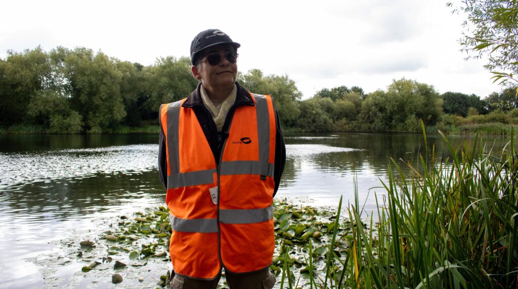 idverde worker in hi-vis vest standing beside a calm lake surrounded by reeds