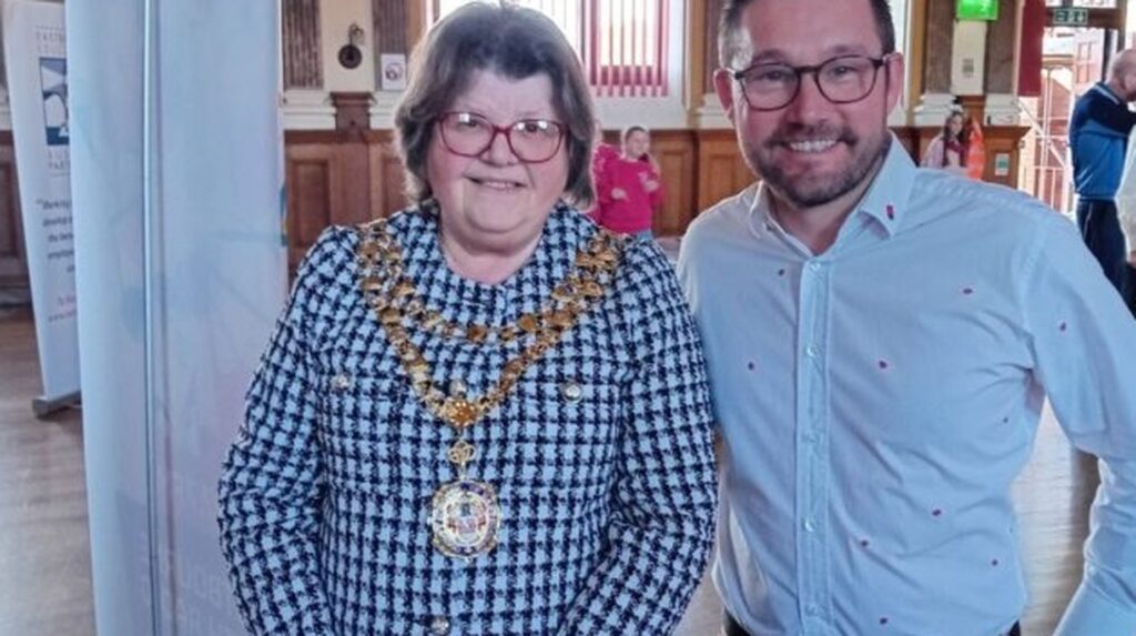 Mayor wearing ceremonial chain poses with man in office at Music in Bloom event