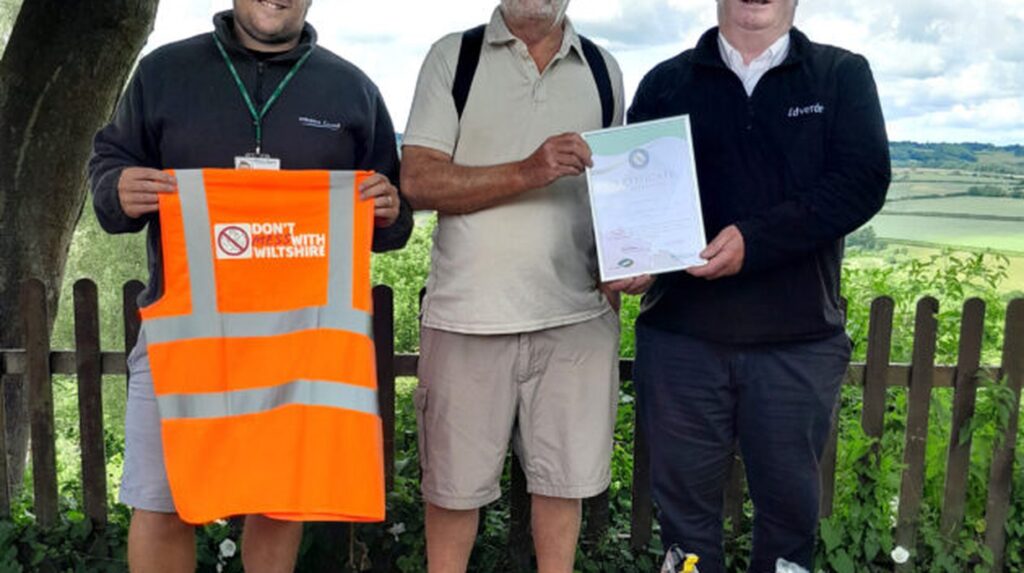 Three men outdoors holding a certificate and hi-vis vest reading 'Don't Mess With Wiltshire'