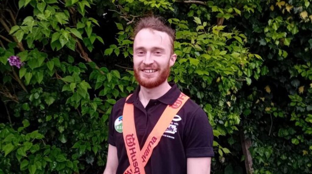 Smiling man with red beard wearing Husqvarna sash over black polo shirt outdoors