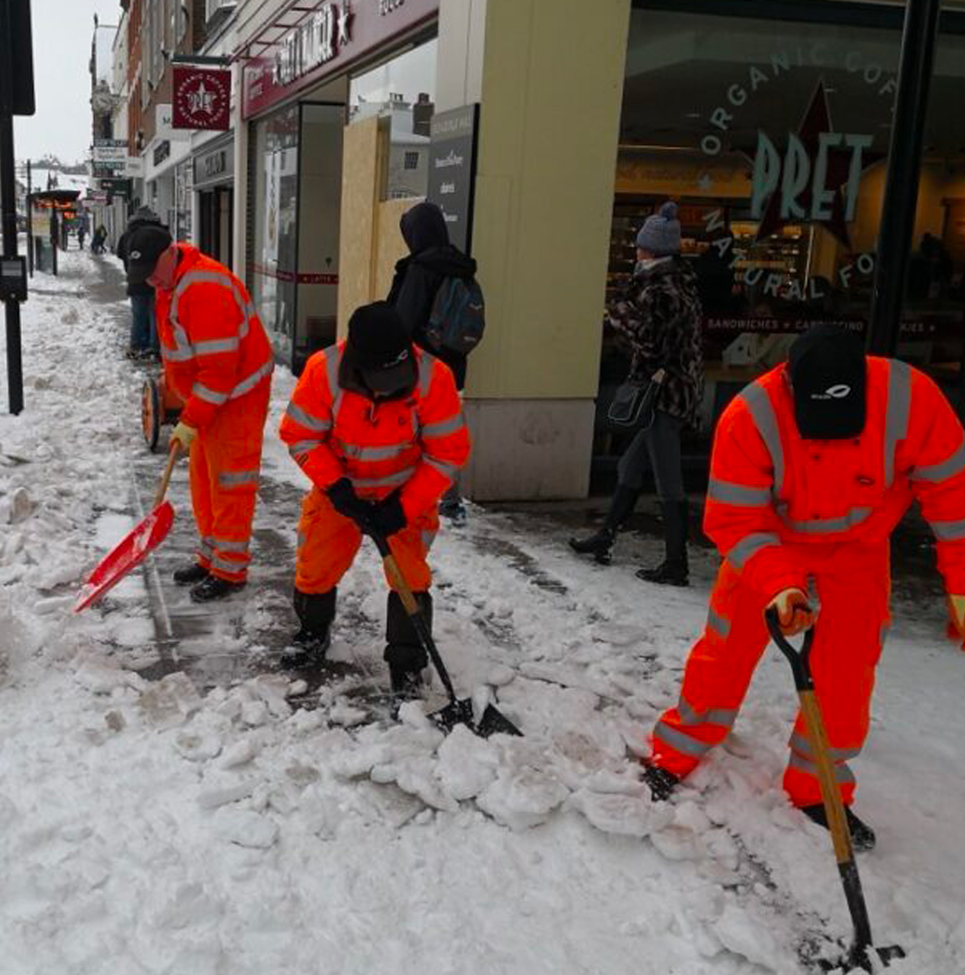 Three idverde operatives in orange high-visibility coveralls manually shovelling snow from a town centre pavement outside a Pret A Manger.