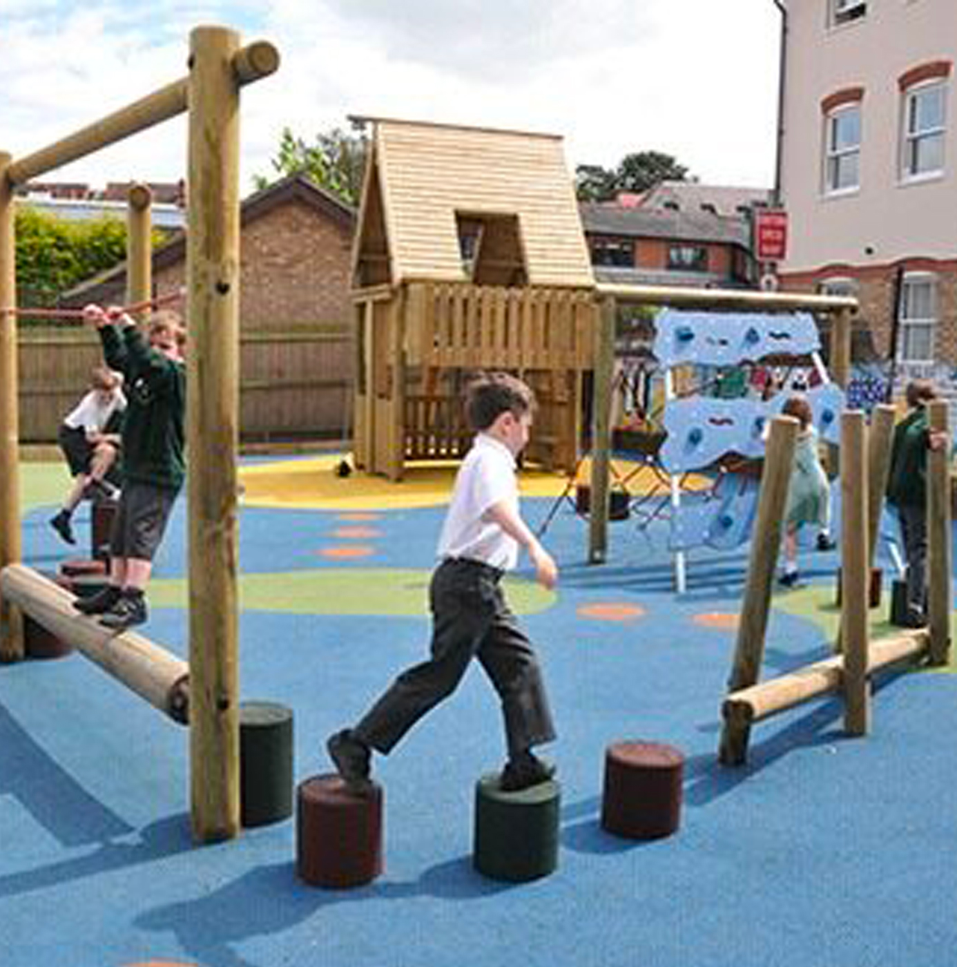 Primary school children playing on a timber and rubber safety surface playground featuring climbing frames, stepping posts, a climbing wall, and a timber playhouse.