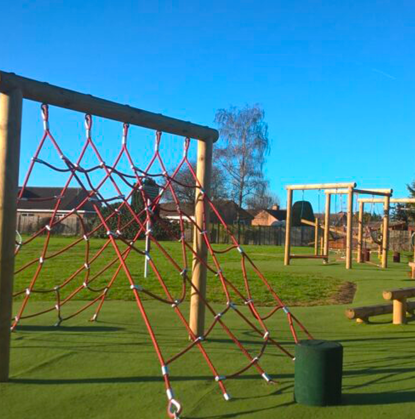A red rope climbing net frame and timber swing set on an artificial grass play area under a clear blue winter sky.