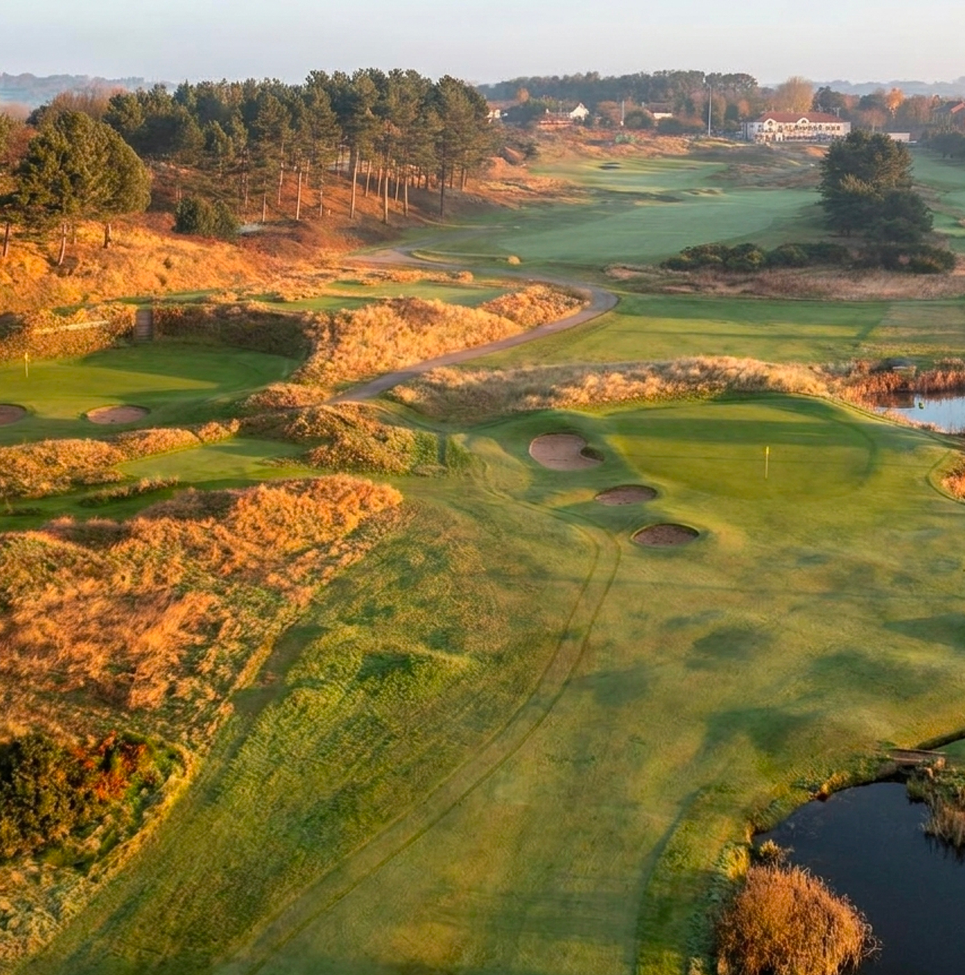 Aerial view of a links golf course at dawn showing manicured fairways, sand bunkers, rough grass areas, water features, and Scots pine tree lines.