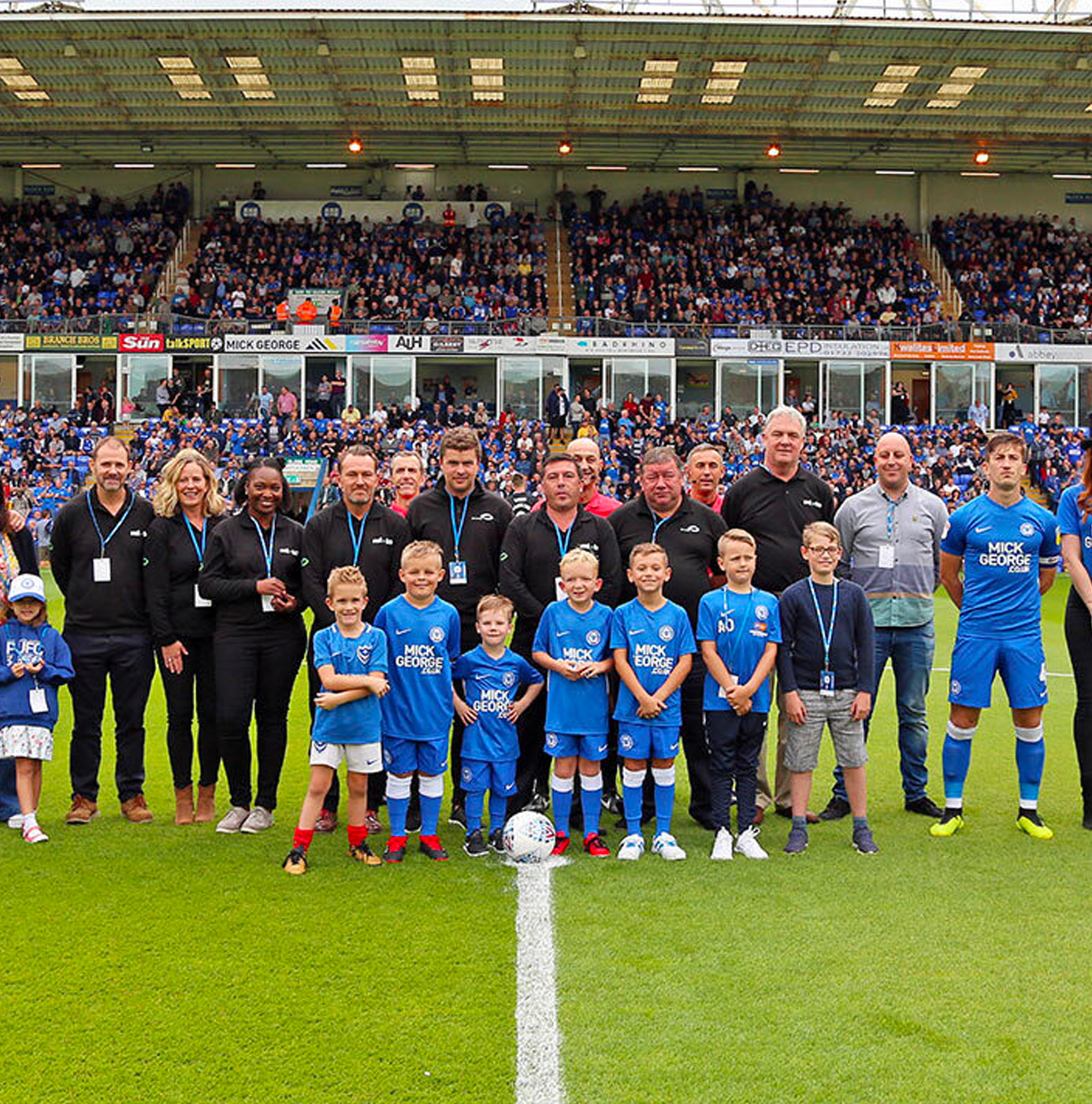 idverde groundscare team and club staff posing on the centre circle of a professional football pitch at the start of a match day, with a full stadium crowd behind.