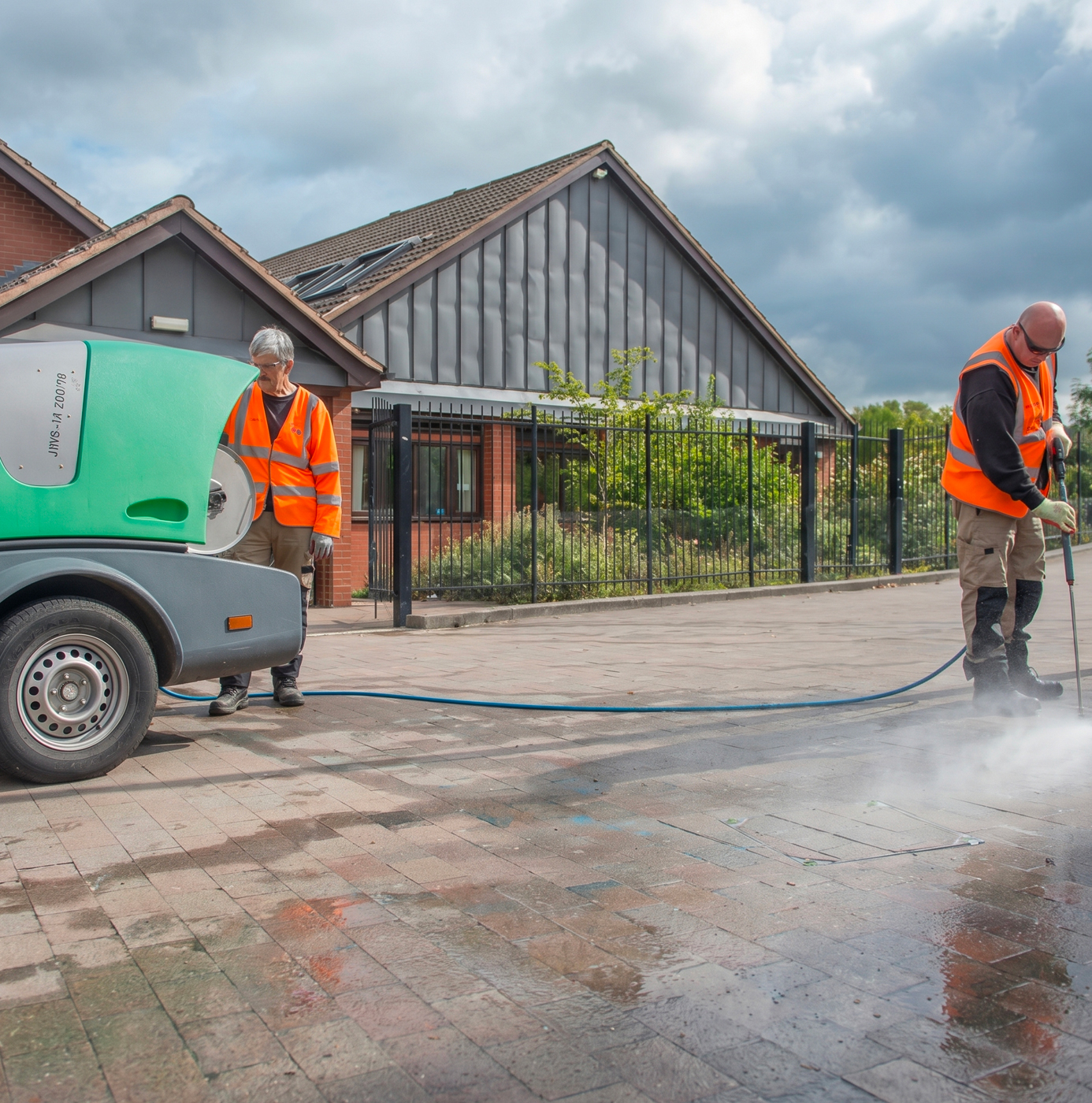 Two idverde operatives in orange high-visibility vests using a hot water pressure washer to clean a block-paved public area beside a residential street.