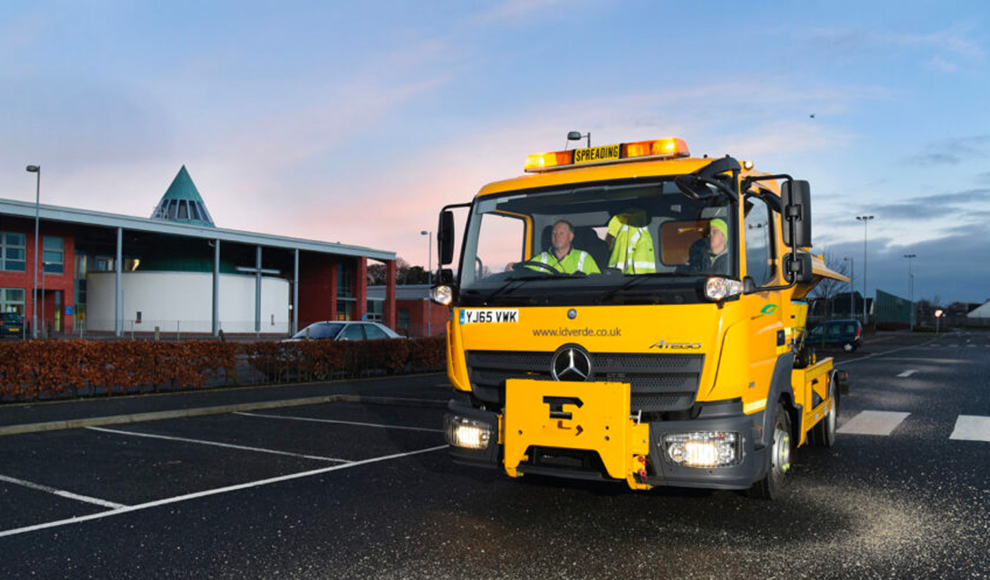 An idverde-branded yellow gritter truck with spreading sign illuminated, crewed by two operatives in high-visibility vests, departing on a pre-dawn winter gritting run.