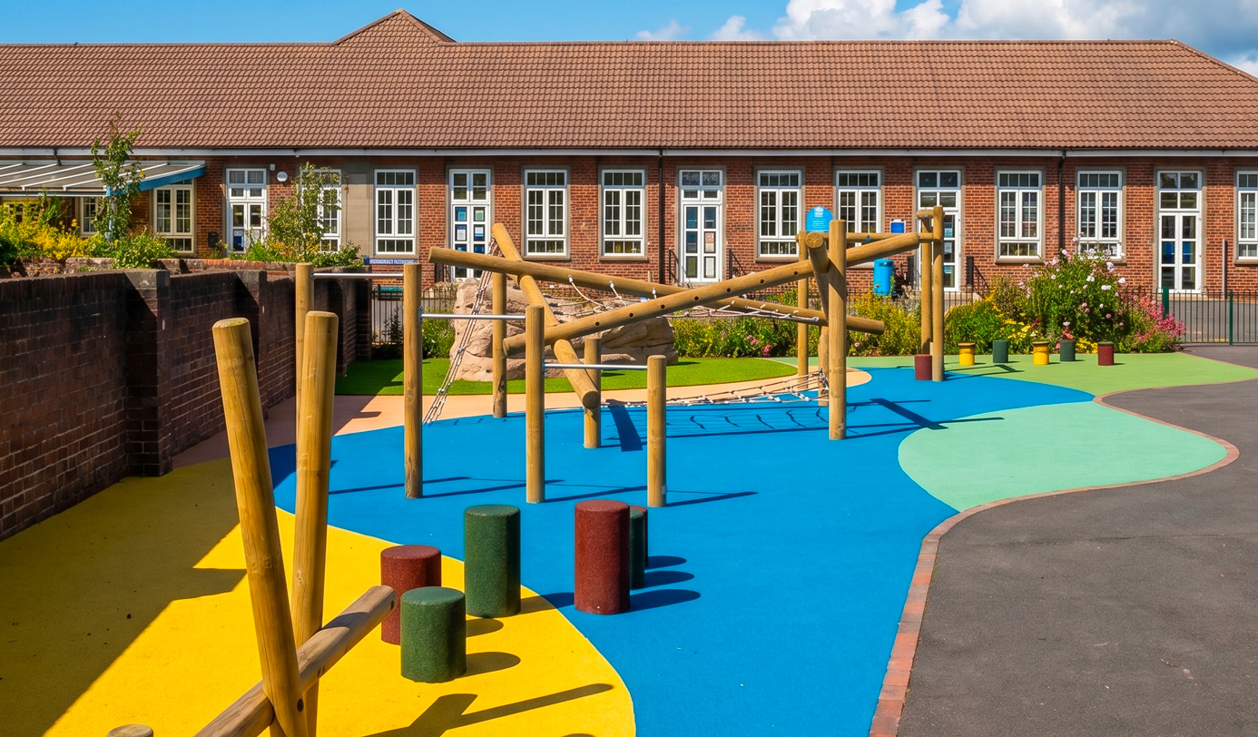 A brightly coloured primary school playground with multi-zone rubberised surfacing, timber climbing frames, stepping posts, and flowering planted borders.