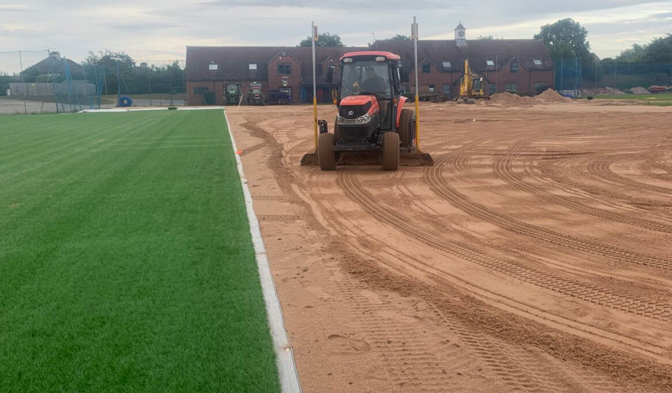 A tractor grading a sand sub-base alongside a completed artificial grass pitch during a multi-pitch sport surface installation.
