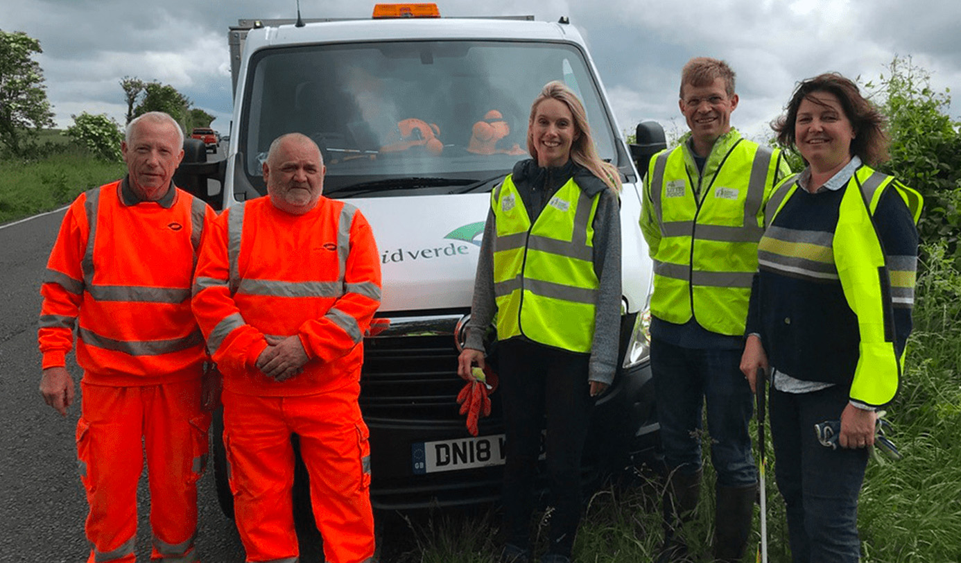 An idverde litter-picking crew and client representatives posing beside an idverde-branded van on a rural road during a highway verge cleaning operation.