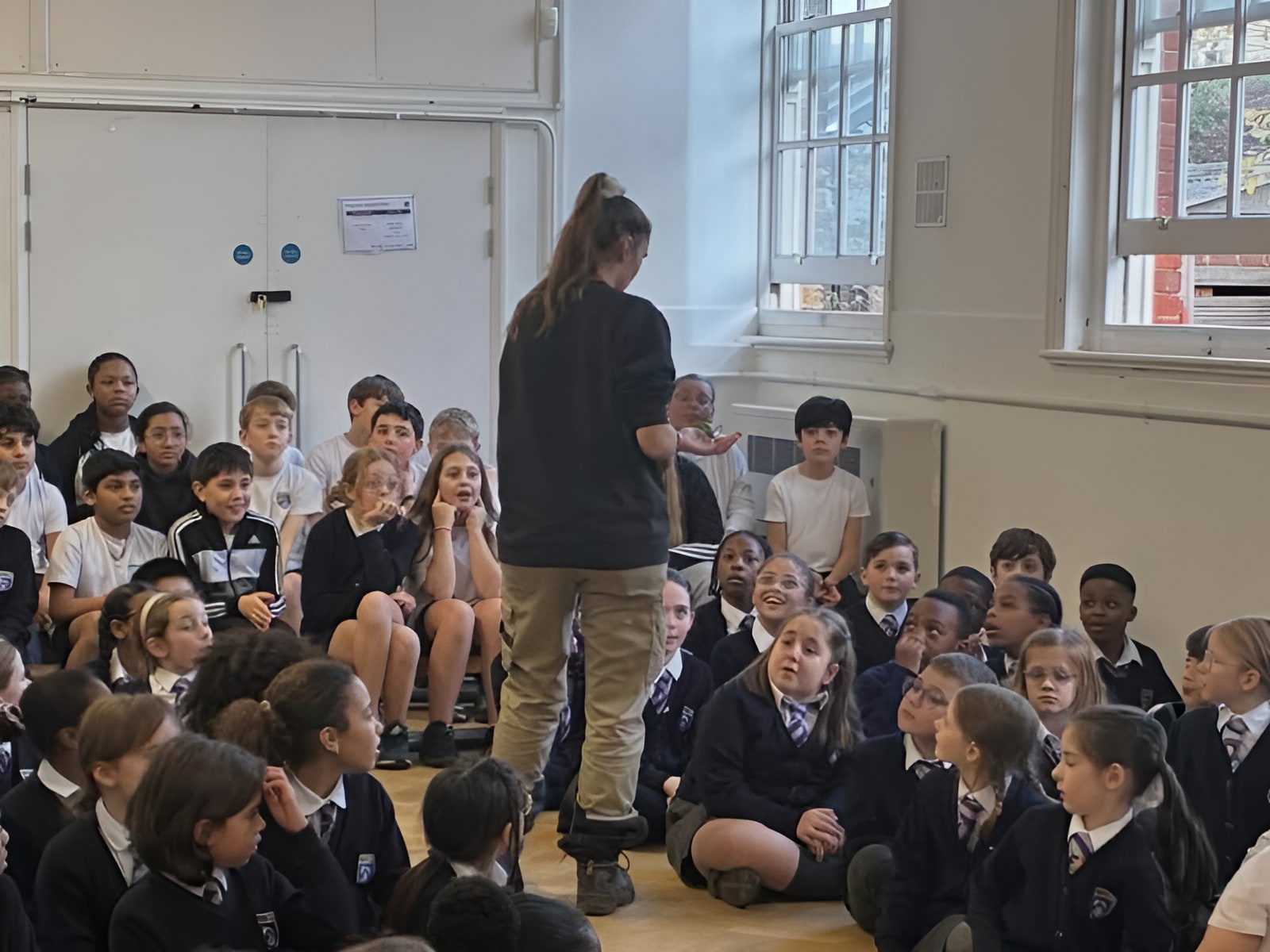 Educator in black top addressing a large group of uniformed school children seated on a hall floor during an assembly.