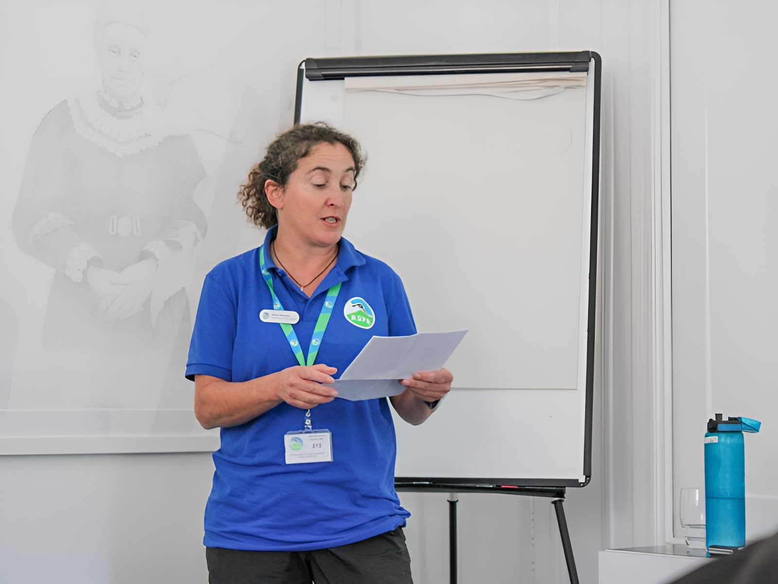 Woman in blue idverde polo shirt presenting from notes at a flipchart during an indoor training session.