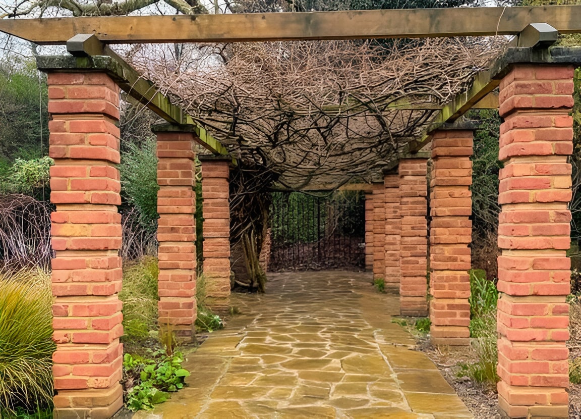 Brick pergola with wooden beams and climbing vines over a stone pathway leading to ornate iron gates